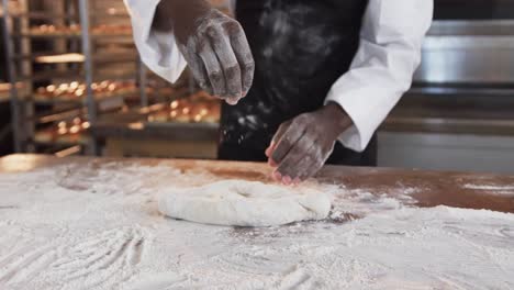 African-american-male-baker-working-in-bakery-kitchen,-pouring-flour-on-dough-in-slow-motion