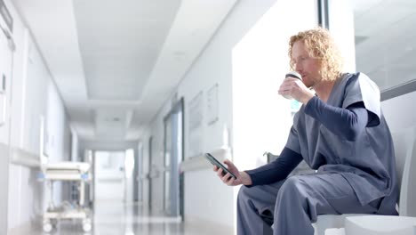 Caucasian-male-doctor-using-smartphone-and-drinking-coffee-in-corridor,-copy-space,-slow-motion