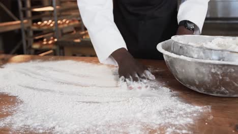 African-american-male-baker-working-in-bakery-kitchen,-spreading-flour-on-counter-in-slow-motion