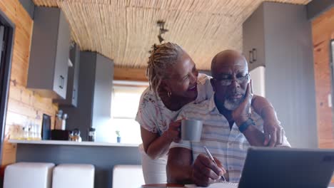 Älteres-Afroamerikanisches-Paar-Umarmt-Und-Erledigt-Papierkram-Mit-Einem-Tablet-Zu-Hause,-Zeitlupe