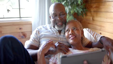 Happy-senior-african-american-couple-using-tablet-lying-on-sofa-at-home,-slow-motion