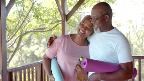 Glückliches-älteres-Afroamerikanisches-Paar,-Das-Matten-Hält-Und-Sich-Auf-Der-Sonnigen-Terrasse-Umarmt,-Zeitlupe