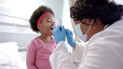African-american-female-doctor-with-face-mask-examining-throat-of-girl-in-hospital-room,-slow-motion