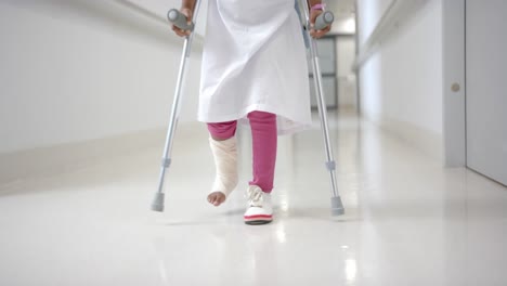 Lowsection-of-african-american-girl-in-hospital-gown-walking-using-crutches-in-corridor,-slow-motion