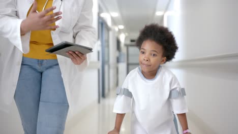 African-american-female-doctor-with-tablet-walking-with-girl-using-crutches-in-corridor,-slow-motion