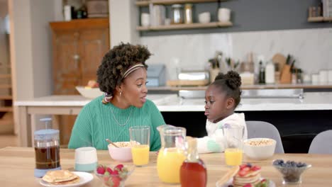 African-american-mother-and-daughter-eating-cereal-with-milk-and-talking-in-kitchen,-slow-motion