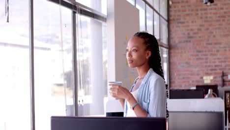 Mujer-De-Negocios-Afroamericana-Informal-Tomando-Café-Y-Mirando-Por-La-Ventana-En-La-Oficina,-Cámara-Lenta