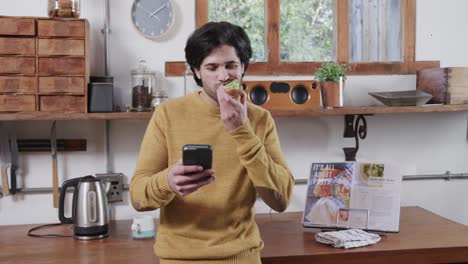 Caucasian-man-eating-avocado-toast-and-using-smartphone-in-kitchen,-slow-motion