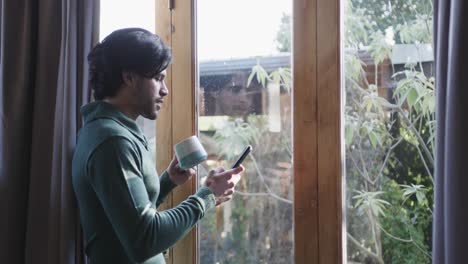 Kaukasischer-Mann-Hält-Eine-Tasse-Kaffee-Mit-Smartphone-Am-Fenster-Zu-Hause,-Kopierraum,-Zeitlupe