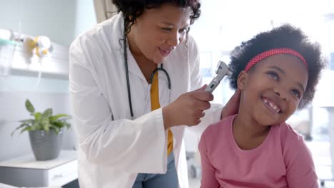 Happy-african-american-female-doctor-examining-ear-of-girl-in-hospital-room,-slow-motion