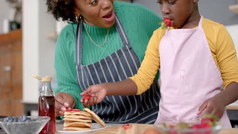 Happy-african-american-mother-and-daughter-decorating-pancakes-with-fruits-in-kitchen,-slow-motion