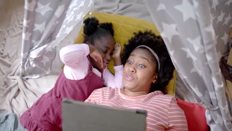 Happy-african-american-mother-and-daughter-lying-in-tent-and-using-tablet-at-home,-slow-motion