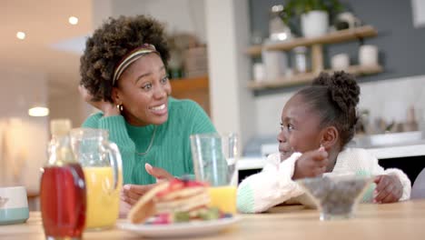 Happy-african-american-mother-and-daughter-eating-cereal-with-milk-in-kitchen,-slow-motion