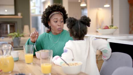 African-american-mother-and-daughter-eating-cereal-with-milk-and-talking-in-kitchen,-slow-motion