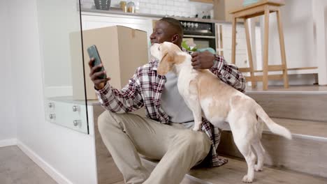 Happy-african-american-man-sitting-on-stairs-using-smartphone,-with-his-pet-dog-at-home,-slow-motion