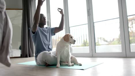 African-american-man-doing-yoga-and-meditating,-with-his-pet-dog-at-home,-slow-motion