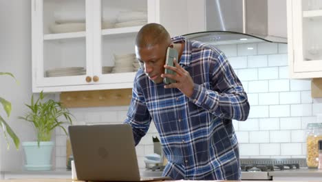 Afroamerikanischer-Mann-Benutzt-Laptop-Und-Spricht-Zu-Hause-In-Der-Küche-Auf-Dem-Smartphone,-Zeitlupe