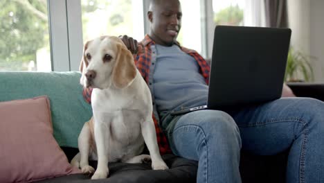 Happy-african-american-man-sitting-on-sofa-using-laptop,-with-his-pet-dog-at-home,-slow-motion