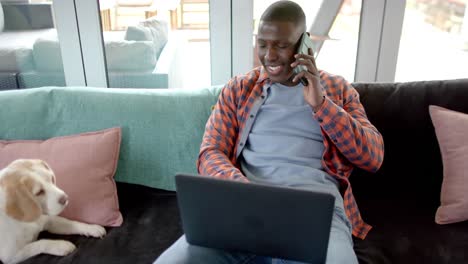 Glücklicher-Afroamerikanischer-Mann-Auf-Smartphone-Und-Laptop-Zu-Hause,-Mit-Seinem-Hund,-Zeitlupe