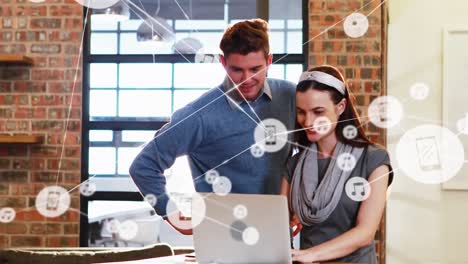Animation-of-network-of-digital-icons-over-caucasian-man-and-woman-discussing-over-laptop-at-office