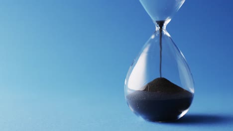 Video-of-close-up-of-hourglass-with-black-sand-pouring-with-copy-space-on-blue-background