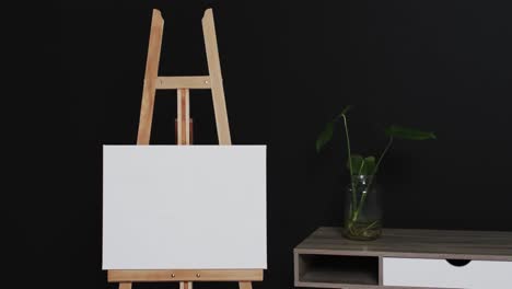 Video-of-white-canvas-sign-on-wooden-easel-with-copy-space-and-plant-on-desk-on-black-background