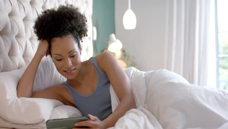 Happy-biracial-woman-lying-in-bed-and-using-smartphone-in-bedroom,-slow-motion