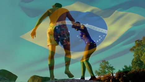 Animation-of-waving-brazil-flag-against-biracial-couple-climbing-rocks-near-the-beach