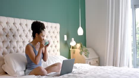 Biracial-woman-sitting-on-bed,-drinking-coffee-and-using-laptop-in-bedroom,-slow-motion