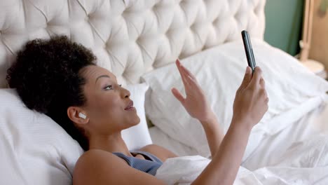 Happy-biracial-woman-with-earphones-making-video-call-using-smartphone-in-bedroom,-slow-motion