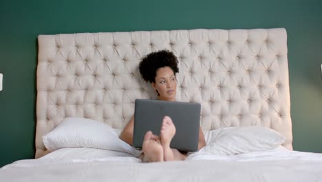 Biracial-woman-sitting-on-bed-and-using-laptop-in-bedroom,-slow-motion