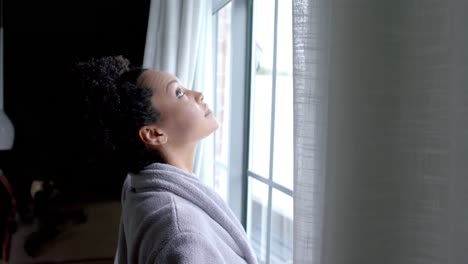 Biracial-woman-in-bathrobe-opening-curtains-in-bedroom,-slow-motion,-copy-space