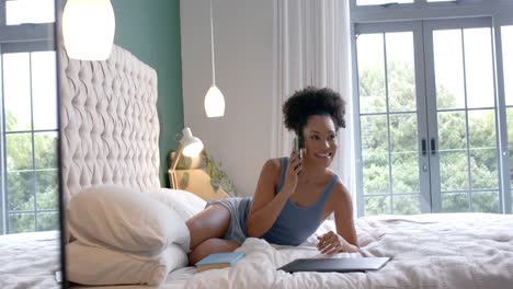 Happy-biracial-woman-lying-on-bed-and-talking-on-smartphone-in-bedroom,-slow-motion