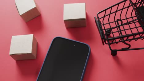 Video-of-smartphone-with-blank-screen,-boxes-and-shopping-trolley-with-copy-space-on-red-background