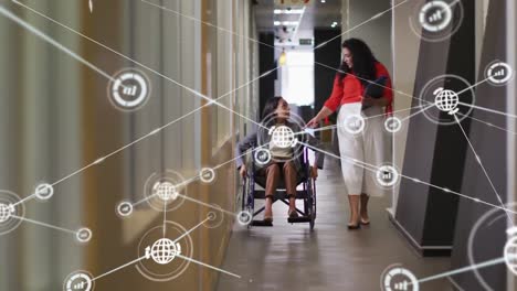 Animation-of-connected-graph-and-globe-icons,-diverse-woman-in-wheelchair-discussing-with-colleague