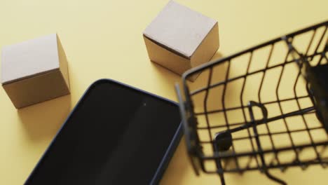 Video-Eines-Smartphones-Mit-Leerem-Bildschirm,-Kartons,-Einkaufswagen-Auf-Gelbem-Hintergrund
