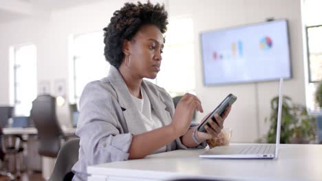 African-american-casual-businesswoman-eating-lunch-and-using-smartphone-in-office,-slow-motion