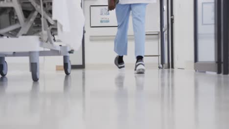 Sección-Baja-De-Un-Médico-Afroamericano-Con-Un-Teléfono-Inteligente-Caminando-En-El-Pasillo-Del-Hospital,-En-Cámara-Lenta