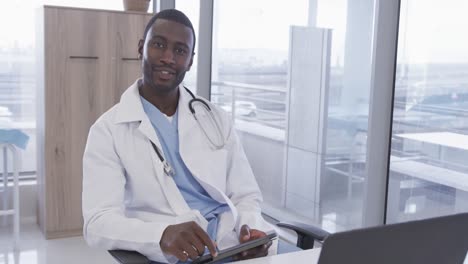 Portrait-of-happy-african-american-male-doctor-using-tablet-in-hospital-office,-slow-motion