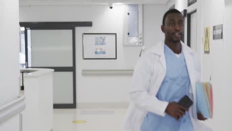 Médico-Afroamericano-Sosteniendo-Un-Teléfono-Inteligente-Y-Corriendo-En-El-Pasillo-Del-Hospital,-Cámara-Lenta