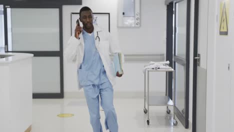 African-american-male-doctor-talking-on-smartphone-and-running-in-hospital-corridor,-slow-motion