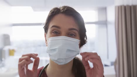 Portrait-of-happy-caucasian-female-doctor-with-face-mask-in-hospital-room,-slow-motion