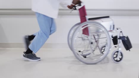 Unterteil-Eines-Afroamerikanischen-Männlichen-Arztes,-Der-Mit-Rollstuhl-Im-Krankenhausflur-Läuft,-Zeitlupe