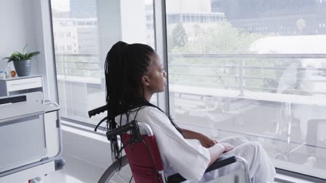 African-american-female-patient-in-wheelchair-looking-out-window-in-hospital-room,-slow-motion