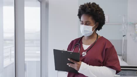 African-american-female-doctor-wearing-face-mask-and-holding-documents-in-hospital-room,-slow-motion