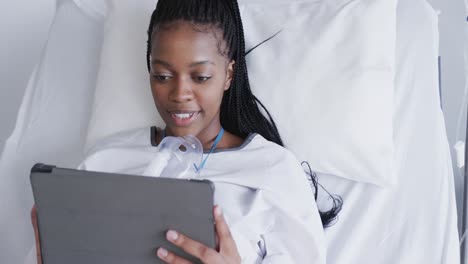 Happy-african-american-female-patient-using-tablet-lying-in-bed-in-hospital-room,-slow-motion