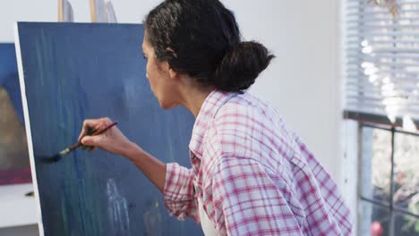 Focused-biracial-female-artist-in-apron-painting-on-canvas-in-studio,-slow-motion