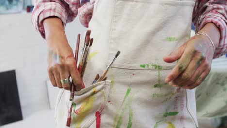 Midsection-of-biracial-female-artist-putting-paint-brushes-in-pocket-of-apron-in-studio,-slow-motion