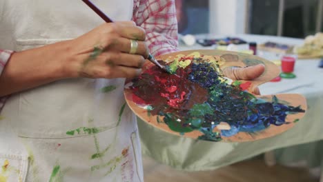 Hands-of-biracial-female-artist-in-apron-mixing-paints-on-palette-using-brush-in-studio,-slow-motion