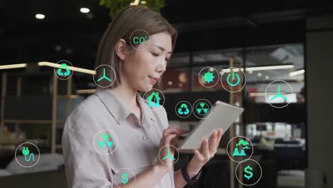 Animation-of-multiple-icons-over-asian-woman-standing-and-scrolling-on-digital-tablet-in-office